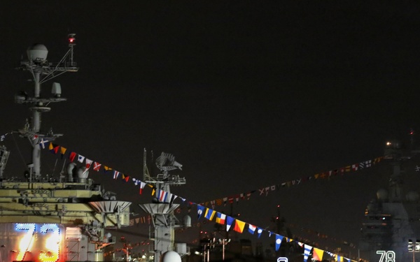 A nighttime photograph of what appears to be two U.S. Navy aircraft carriers docked at a naval facility. The vessels display hull numbers 73 and 78, decorated with strings of colorful signal flags (dress ship bunting) suggesting a ceremonial occasion. The scene is dimly lit with artificial lighting illuminating the ships' superstructures and hull numbers.