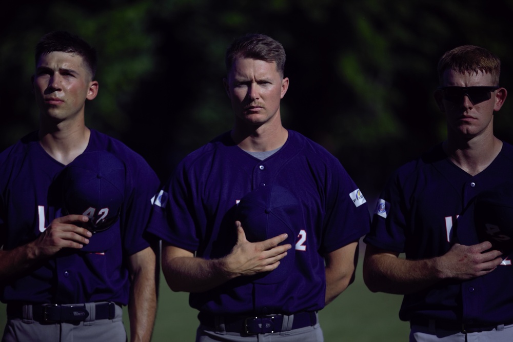 Three baseball players in matching purple/navy uniforms stand in a line with their right hands placed over their hearts, suggesting they are participating in a national anthem ceremony. The players appear solemn and attentive, facing forward in what appears to be a pre-game ritual. The background shows dark green foliage, and the lighting creates a dramatic contrast on the players' faces.