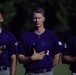 Three baseball players in matching purple/navy uniforms stand in a line with their right hands placed over their hearts, suggesting they are participating in a national anthem ceremony. The players appear solemn and attentive, facing forward in what appears to be a pre-game ritual. The background shows dark green foliage, and the lighting creates a dramatic contrast on the players' faces.