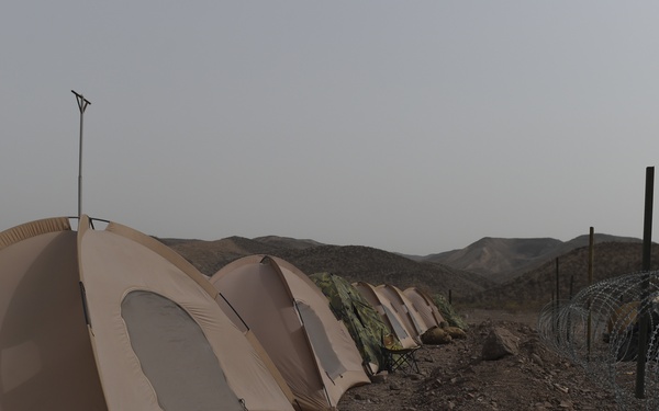 A row of tan/beige military-style dome tents is set up in an arid, rocky desert landscape with mountains visible in the background. The camp is secured with concertina (razor) wire coiled around posts on the right side of the image. The environment appears harsh and dry, consistent with a forward operating base or field encampment.