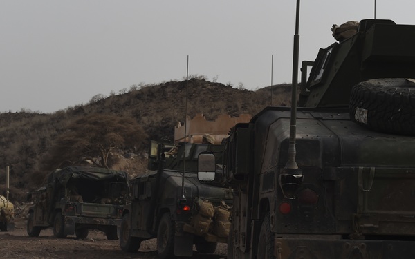 A military convoy of tactical vehicles is staged or moving along a dirt road in an arid, mountainous terrain. The vehicles appear to be a mix of HMMWVs (Humvees) and a larger armored vehicle in the foreground, consistent with U.S. or coalition military operations. The environment is dry and barren with low hills in the background under an overcast sky.
