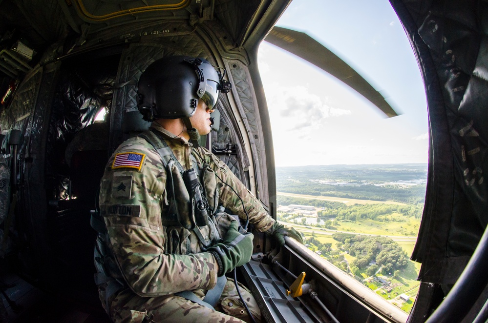 A U.S. Army soldier wearing multicam combat uniform and a flight helmet with communications gear sits inside a military helicopter, looking out through a large open window at a green, rural landscape below. The soldier has a U.S. flag patch on the left shoulder and rank insignia visible on the uniform. The wide-angle fisheye lens captures both the interior of the aircraft and the aerial view of the terrain.