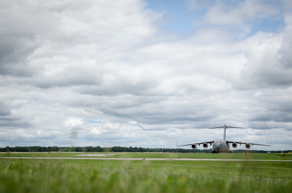 A large military cargo aircraft, consistent with a C-17 Globemaster III, is positioned on or near a runway at a military airfield. The aircraft is viewed from a low ground-level perspective, emphasizing the expansive green grass in the foreground and a heavily overcast sky above. The setting appears to be a well-maintained airfield surrounded by tree lines in the background.