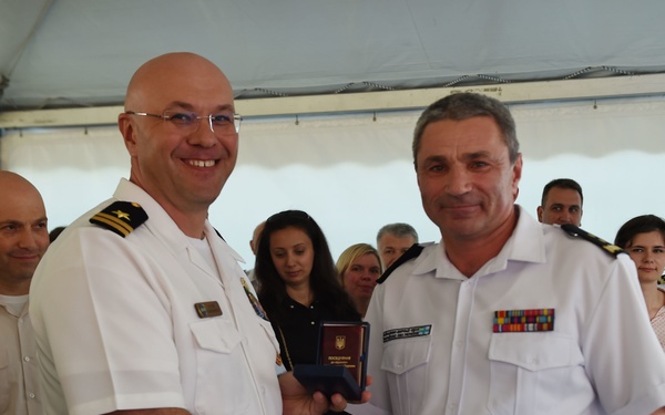 Two naval officers in white dress uniforms are shaking hands during what appears to be a formal military ceremony. One officer is presenting the other with a small booklet or credential bearing what appears to be a Ukrainian trident emblem. The event is taking place under a white tent with a group of military personnel and civilians observing in the background.