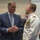 Marine Barracks Washington; EveningParade July 14, 2017 Two men are engaged in conversation at what appears to be a formal reception or ceremony. One man is wearing a civilian blue suit with a tie, while the other is dressed in a white U.S. Navy dress uniform adorned with numerous medals, ribbons, and gold insignia indicating high rank. Several other attendees are visible in the background.