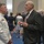 Marine Barracks Washington; EveningParade July 14, 2017 A formal reception or military ceremony is taking place in what appears to be a government or institutional building with a blue carpet. Two men are prominently featured in conversation: one wearing a white U.S. Navy dress uniform with multiple ribbons and gold insignia, and another in a dark business suit holding a glass. The background shows numerous other attendees in formal attire mingling in the space.