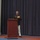 Marine Barracks Washington; EveningParade July 14, 2017 A military officer in dress uniform stands at a burgundy podium holding a microphone, appearing to deliver a speech or address. The individual is wearing what appears to be a U.S. Marine Corps dress blue uniform with multiple rows of service ribbons and medals. The setting appears to be a formal indoor ceremony or event with dark blue curtains as a backdrop.