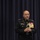 Marine Barracks Washington; EveningParade July 14, 2017 A senior U.S. Marine Corps officer in dress blues uniform is speaking at a formal event, holding a microphone. He is adorned with numerous medals, ribbons, and insignia indicating very high rank, consistent with a general officer. He stands in front of a dark blue curtain backdrop on what appears to be a stage.