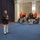 Marine Barracks Washington; EveningParade July 14, 2017 A formal military ceremony or reception is taking place in an elegant indoor venue with blue carpeting and arched architectural details. A senior Marine Corps officer in dress blues with extensive medal decorations appears to be addressing a group that includes Navy officers in white dress uniforms and musicians in red ceremonial uniforms. The setting suggests a high-level official function, likely involving multiple branches of the U.S. military.