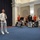 Marine Barracks Washington; EveningParade July 14, 2017 A formal military ceremony or reception is taking place in an elegant hall with classical architectural features. A senior officer in white dress uniform speaks into a microphone while musicians in red ceremonial uniforms and officers in black dress uniforms stand nearby. Civilians are also present, suggesting this is a mixed military-civilian event.