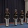 Marine Barracks Washington; EveningParade July 14, 2017 Six United States Marines in dress blue uniform are standing at attention in a formal ceremonial formation. They are holding rifles at their sides and are positioned in two diagonal columns on a blue floor in front of a dark navy curtain backdrop. The scene appears to be an indoor military ceremony or parade drill.