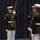 Marine Barracks Washington; EveningParade July 14, 2017 Three United States Marines in dress blue uniform are participating in a formal ceremonial drill or parade. The Marines are holding rifles in a precise ceremonial position, consistent with a Silent Drill Platoon or honor guard performance. The setting appears to be an indoor venue with a dark blue curtain backdrop.