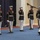 Marine Barracks Washington; EveningParade July 14, 2017 A group of U.S. Marine Corps personnel in dress blue uniforms are performing an indoor ceremonial drill routine. At least one Marine is executing a rifle toss or spin maneuver, with a rifle visibly airborne above head level. The performance is taking place on a blue carpeted floor in what appears to be a formal indoor venue.