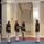 Marine Barracks Washington; EveningParade July 14, 2017 A group of U.S. Marines in dress blues uniform are performing a silent drill or ceremonial rifle exhibition indoors. Two rifles are visibly airborne, having been tossed upward, while the Marines stand at attention or track the rifles with their gaze. The setting appears to be a formal indoor venue with decorative columns and arched alcoves.