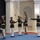 Marine Barracks Washington; EveningParade July 14, 2017 A group of U.S. Marine Corps Silent Drill Platoon members are performing an indoor ceremonial rifle drill routine. Several Marines in dress blues with white trousers and white gloves are executing precise rifle tosses and maneuvers. One Marine on the right appears to be in a command or observing role with arm extended, while two others in the background stand at attention.