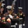 Marine Barracks Washington; EveningParade July 14, 2017 The image shows United States Marine Corps personnel in dress blue uniforms performing a ceremonial drill or silent drill platoon routine. Multiple Marines are holding rifles at precise angles while wearing their formal dress uniforms with white covers, white gloves, and white belts. The setting appears to be an indoor venue with a dark blue curtain backdrop.
