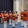 Marine Barracks Washington; EveningParade July 14, 2017 A military band, identifiable as the United States Marine Corps Band based on their distinctive scarlet and white dress uniforms, performs indoors in a formal ceremonial setting. The musicians are playing silver trumpets/bugles while arranged in formation on a blue carpeted floor. A conductor or drum major figure is visible facing the ensemble, distinguished by additional gold braiding on their uniform.