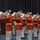 Marine Barracks Washington; EveningParade July 14, 2017 A military marching band, identifiable as the United States Marine Corps Band based on their distinctive red tunics and white trousers, performs in formation. The musicians are playing large brass instruments, likely euphoniums or marching baritones, while wearing formal dress uniforms with white gloves and white peaked caps. The performance appears to be taking place indoors on a stage or ceremonial hall with a dark blue curtain backdrop.