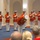 Marine Barracks Washington; EveningParade July 14, 2017 A military band performance is taking place in an indoor venue, with musicians dressed in distinctive red and white dress uniforms holding silver trumpets/bugles. A central figure, likely the conductor or drum major, is bowing toward the audience in the foreground. The setting appears to be a formal ceremonial hall with arched architectural details.