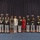 Marine Barracks Washington; EveningParade July 14, 2017 A formal military group photograph featuring United States Marine Corps personnel in dress uniforms alongside two civilian women and a bulldog. The group is posed in front of a dark blue curtain backdrop on a blue-carpeted stage or ceremonial hall. The presence of medals, white dress uniforms, and a regimental mascot (bulldog) suggests a formal ceremony or official event.