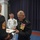 Marine Barracks Washington; EveningParade July 14, 2017 A formal military ceremony is taking place indoors, where a high-ranking Marine Corps general in dress blues is signing an official document presented by a Marine Staff Sergeant. A Navy officer in dress whites observes smiling in the background. The setting appears to be an official ceremonial hall with blue curtains and a red and blue floor.