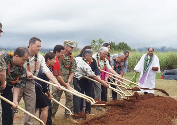 A groundbreaking ceremony is taking place outdoors in what appears to be Hawaii, indicated by the traditional lei garlands worn by participants. A group of approximately 12-14 individuals, including military personnel in camouflage uniforms, civilians, and a clergy member in white robes, are simultaneously shoveling red soil with ceremonial gold-handled shovels. The overcast sky, lush green vegetation, and red volcanic soil are consistent with a Hawaiian setting.