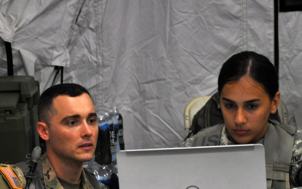 Two U.S. Army soldiers in military uniforms are seated at a white table working together on a Dell laptop computer. The setting appears to be a field operations tent or temporary command post, indicated by the white fabric walls and military equipment visible in the background. One soldier is operating the mouse while both appear focused on the laptop screen.