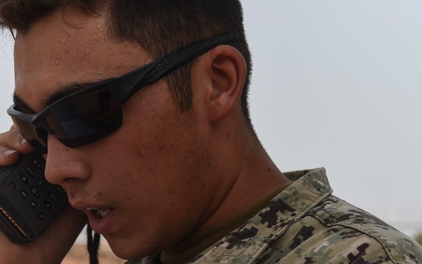 A U.S. Navy service member is shown in close-up, speaking into a handheld radio device. The individual is wearing Navy Working Uniform (NWU) Type III camouflage and tactical sunglasses, suggesting an operational or deployed environment. The arid, desert-like background indicates a forward operating location.