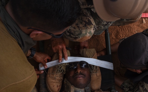 Military personnel in camouflage uniforms are providing tactical combat casualty care (TCCC) to a downed service member on reddish-brown dirt ground. The casualty is wearing a cervical collar and has simulated blood/wounds visible on his arm and shirt, suggesting this is a training exercise. Multiple responders are applying a head immobilization strap and attending to the patient simultaneously.