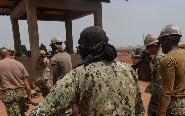 Military personnel in a arid, dusty outdoor environment are engaged in what appears to be a training or operational exercise involving casualty evacuation. Several individuals wearing U.S. Navy digital camouflage (NWU Type III) and tan/khaki uniforms are carrying a litter (stretcher) toward a covered structure. White construction-style hard hats with green cross markings are visible on several personnel, suggesting a medical or engineering context.