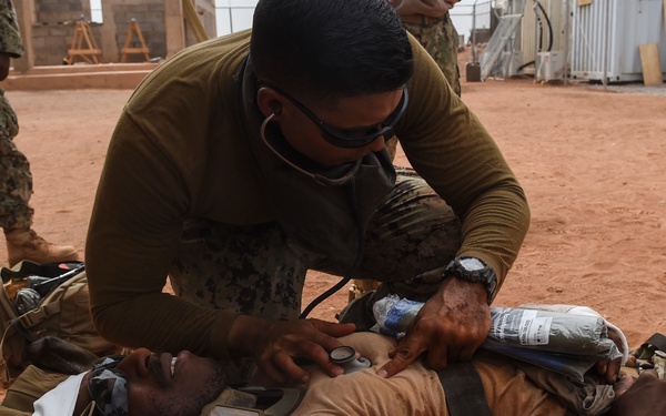 A military medic wearing protective goggles and an olive-colored uniform is providing medical treatment to a casualty lying on a stretcher on red dirt ground. The casualty is wearing camouflage uniform and has visible simulated wounds, a white bandage on their head, and appears to be receiving airway or neck assessment. The setting appears to be a forward operating base or military training compound, with other uniformed personnel visible in the background.