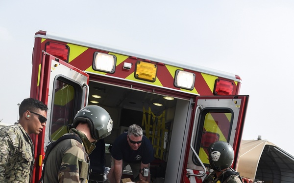 A medical emergency response scene showing multiple personnel transferring a patient on a stretcher into or out of a red and yellow ambulance on what appears to be a military airfield tarmac. Personnel include individuals in French camouflage uniforms with helmets, civilian medical staff, and U.S. Navy personnel. The back of one civilian's shirt reads 'CAMP... &amp; EMERGENCY... Djibouti,' suggesting this is located at Camp Lemonnier or a similar installation in Djibouti.