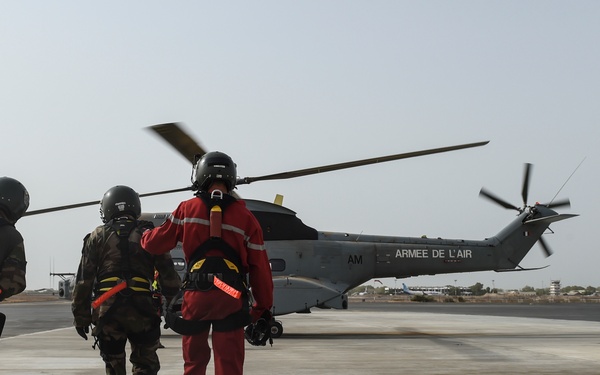 Three personnel wearing helmets and flight gear are walking toward a French Air Force (Armée de l'Air) helicopter on a tarmac. The helicopter's rotors are spinning, indicating it is active and preparing for or returning from a mission. The scene appears to take place at a military airfield in an arid environment.
