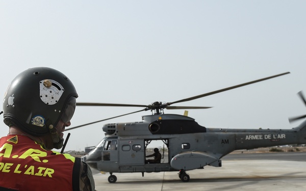 A French Air Force (Armée de l'Air) SAR (Search and Rescue) personnel stands on a tarmac observing a military helicopter with rotors spinning. The individual is wearing a high-visibility yellow/red vest and a flight helmet with communications equipment. The helicopter, marked 'ARMEE DE L'AIR', appears to be preparing for takeoff or has just landed.