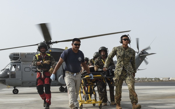 A medical evacuation scene on a military airfield tarmac showing personnel transporting a patient on a yellow stretcher away from a military helicopter. The team consists of mixed personnel including what appears to be a US Navy sailor in camouflage uniform, a civilian or paramedic in a blue shirt, and flight crew members in helmets and flight suits. The helicopter in the background has its rotors spinning, indicating a recent or ongoing MEDEVAC operation.