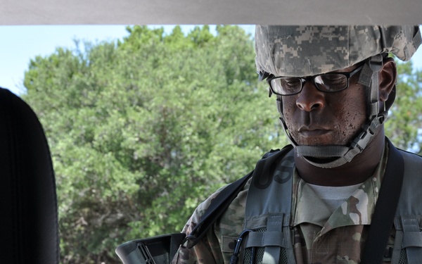 A U.S. Army soldier in full combat gear is leaning into or standing beside a vehicle, appearing to inspect or handle equipment. The soldier is wearing Army Combat Uniform (ACU/Multicam pattern), a tactical vest, ballistic helmet, and eyeglasses. The setting appears to be an outdoor training environment with trees and open terrain visible in the background.