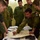 A group of military aviators in olive/green flight suits are gathered around a table conducting what appears to be a mission or flight planning briefing. Multiple nationalities are represented, with visible patches including a Canadian flag and a US flag on flight suits. One individual is pointing to a aeronautical navigation chart while others observe attentively.