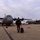 A military personnel member in olive/green flight suit walks across a tarmac toward a large gray turboprop military transport aircraft, pulling a rolling suitcase and carrying a backpack. Two C-130 Hercules-type aircraft are visible on the flight line, with the right aircraft bearing a Canadian flag marking. The scene appears to be at a military air base under overcast skies.