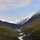 A sweeping mountain valley view captured through what appears to be a vehicle or train window, showing a U-shaped glacial valley flanked by dense coniferous forest. Snow-capped jagged mountain peaks dominate the background, with a partially frozen or snow-covered valley floor visible below. The scene is consistent with the Canadian Rockies landscape.