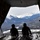 Two personnel in flight helmets sit on the open rear ramp of a military cargo aircraft, overlooking a snow-capped mountain range and forested valley below. The image is taken from inside the aircraft looking outward through the open ramp door. The dramatic silhouette of the figures contrasts with the bright, expansive mountain landscape visible beyond.