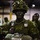 A soldier in full combat gear stands in the foreground of what appears to be a military staging area or hangar. The soldier is wearing Canadian CADPAT (Canadian Disruptive Pattern) camouflage uniform, has face camouflage paint applied, a helmet adorned with foliage, and parachute rigging equipment. Additional soldiers in similar gear are visible in the blurred background.