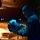 A U.S. military aviator or aircrew member is inspecting a flight helmet at night on what appears to be a flight line or aircraft hangar apron. The individual is wearing a blue flight suit with a U.S. flag patch on the left shoulder. The scene is dramatically lit with blue ambient lighting contrasting against warm orange background illumination.