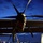 Two military personnel are conducting pre-flight or maintenance checks on a large military aircraft at dusk or dawn on a wet tarmac. The image is dominated by the rear view of a large multi-blade propeller assembly with a red-capped engine inlet visible. One technician is on a blue maintenance stand while another is positioned higher near the engine nacelle.