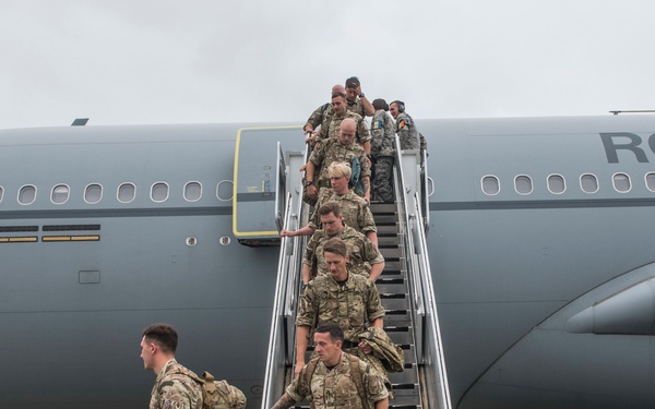 Military personnel in MultiCam/MTP camouflage uniforms are disembarking from a large grey military transport aircraft marked 'ROYAL' (consistent with Royal Air Force markings) via a mobile staircase onto a tarmac. The aircraft displays a RAF roundel on its fuselage and appears to be an Airbus A330 Voyager or similar wide-body transport. The scene suggests an arrival at a military airfield.