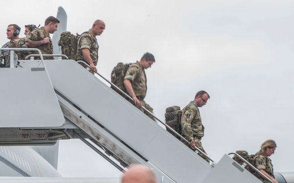 A large group of military personnel in camouflage uniforms are disembarking from a military transport aircraft via two stairways. The soldiers are carrying backpacks and personal gear as they descend onto the tarmac. The scene appears to depict a troop arrival or deployment operation at an airfield.