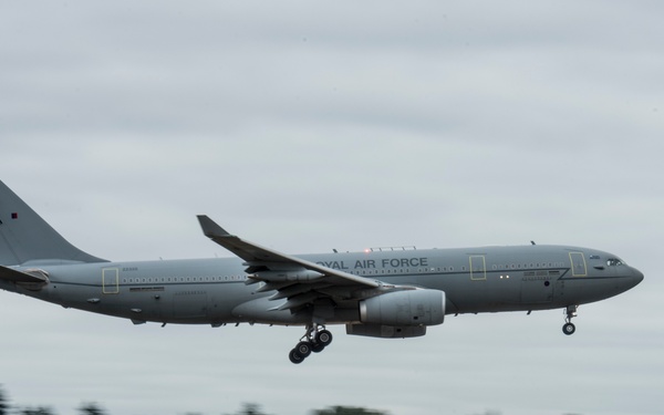 A Royal Air Force (RAF) Airbus A330 Voyager tanker/transport aircraft is captured on approach to landing with its landing gear deployed. The aircraft is painted in an overall grey military livery with 'ROYAL AIR FORCE' markings visible on the fuselage. The background shows a tree line and airfield infrastructure, with motion blur suggesting a panning camera technique.