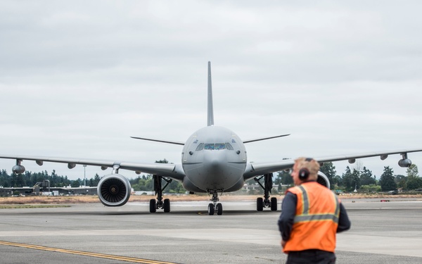 A large gray military or government aircraft, consistent with a KC-46 Pegasus or similar wide-body tanker/transport, is taxiing on a runway apron toward the camera. A ground crew member wearing an orange high-visibility vest and headset stands facing the aircraft, likely providing marshaling guidance. The scene appears to be at a military or joint-use airfield under overcast skies.