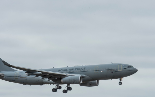 A Royal Air Force aircraft, identifiable as an Airbus A330 MRTT (Multi Role Tanker Transport), is captured on approach to landing with its landing gear fully deployed. The aircraft is painted in a standard RAF grey livery with 'ROYAL AIR FORCE' text clearly visible on the fuselage. The image was taken from ground level near an airfield, with trees and low buildings visible in the background under overcast skies.