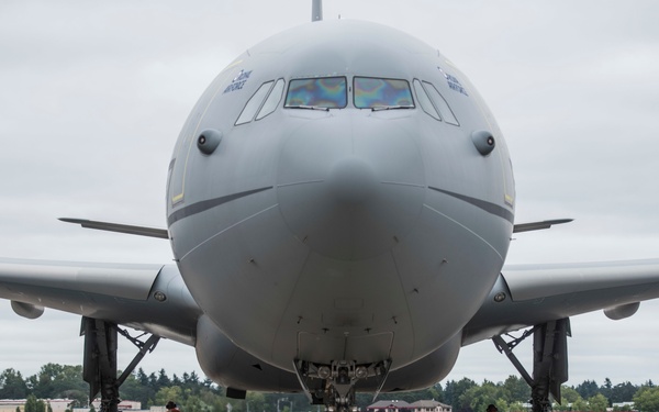 A large military-style gray aircraft is parked on a tarmac, viewed head-on from the front. Two ground crew members wearing orange high-visibility vests are positioned near the main landing gear on each side. The aircraft appears to be a Boeing KC-46 or similar wide-body tanker/transport aircraft based on its configuration and gray livery.