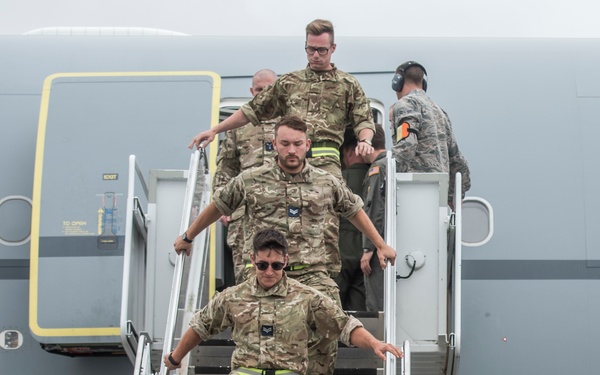 Military personnel in MultiCam/MTP camouflage uniforms are descending the airstairs of a large military or charter aircraft. The group consists of approximately four to five individuals, with one person in a different uniform (ABU/grey pattern) visible at the top of the stairs. The setting appears to be a military airfield or airport tarmac.
